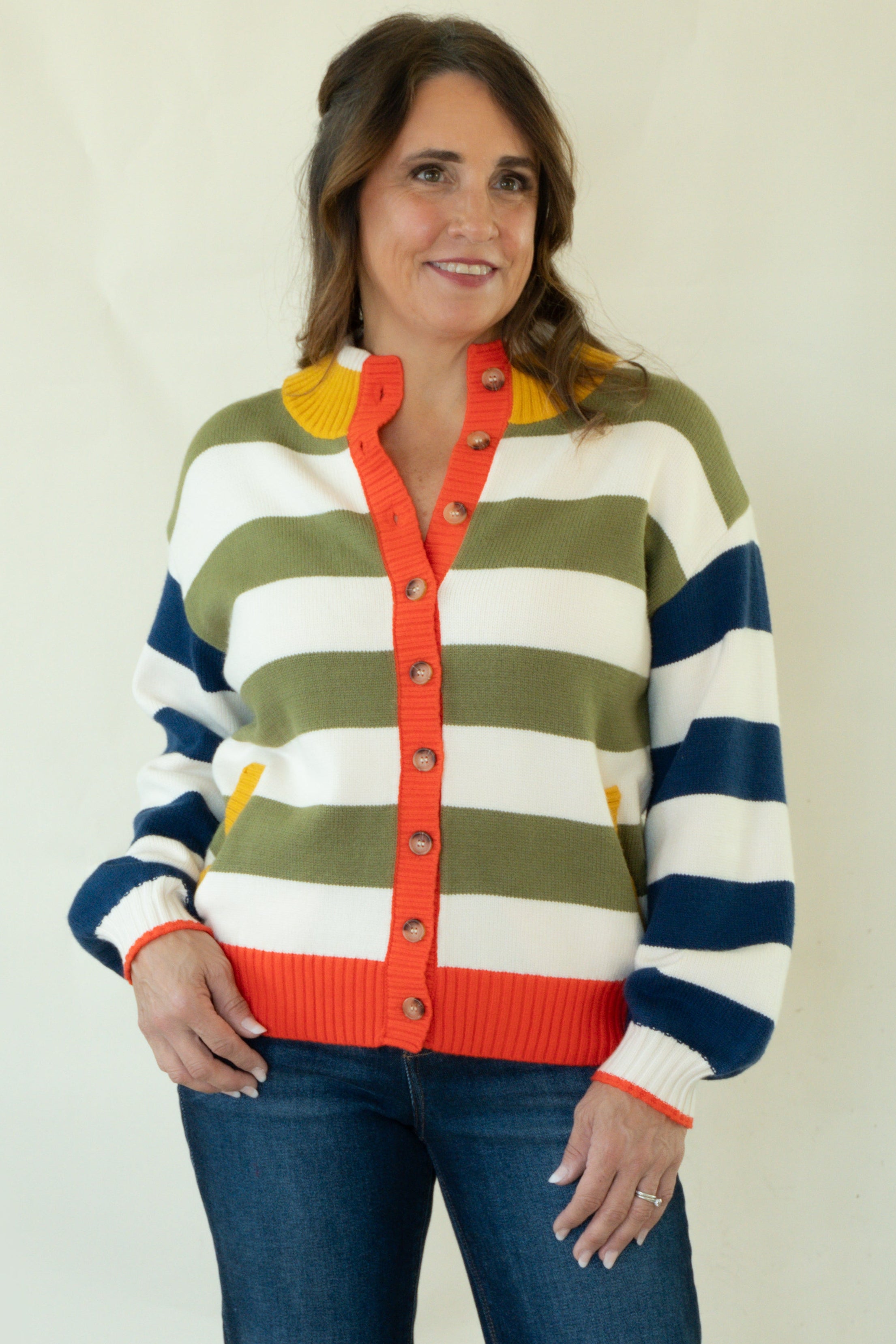 A woman stands barefoot against a plain off-white studio background wearing a striped knit cardigan with blue, olive green, white, and orange horizontal bands, paired with dark blue jeans.