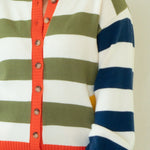 A woman stands barefoot against a plain off-white studio background, wearing a colorful striped cardigan with blue jeans.