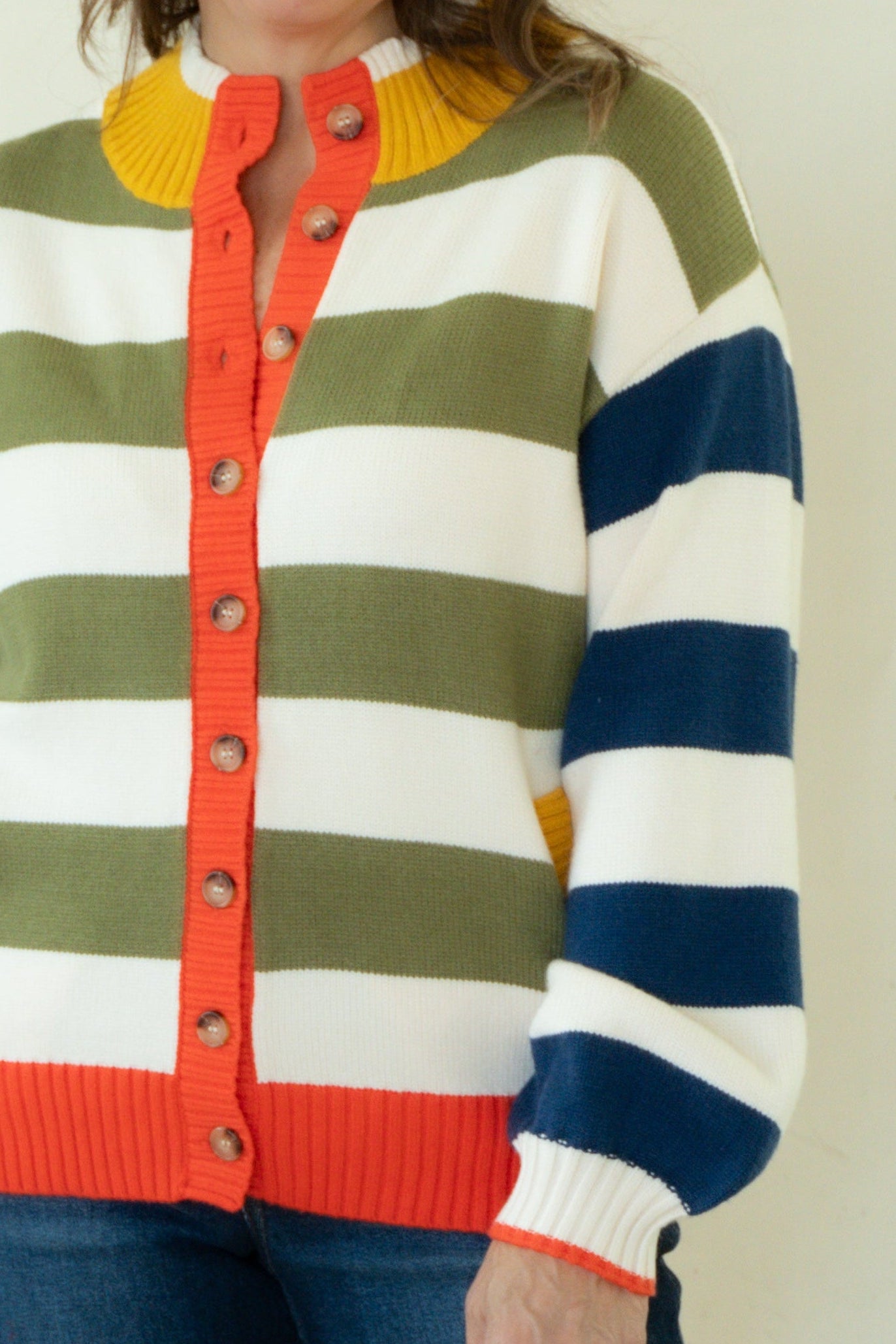A woman stands barefoot against a plain off-white studio background, wearing a colorful striped cardigan with blue jeans.
