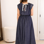 Woman in a long denim dress standing in a room with a mirror and furniture.