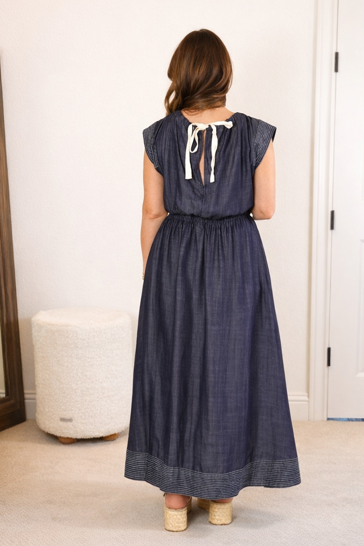 Woman in a long denim dress standing in a room with a mirror and furniture.