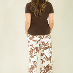 A woman viewed from behind wearing a dark brown short-sleeve t-shirt and wide-leg pants with a brown and white cow-print pattern, standing against a plain light beige studio background.