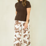 A woman modeling a brown short-sleeve mock neck top and white wide-leg pants with a brown abstract floral pattern, standing against a plain light beige studio background.