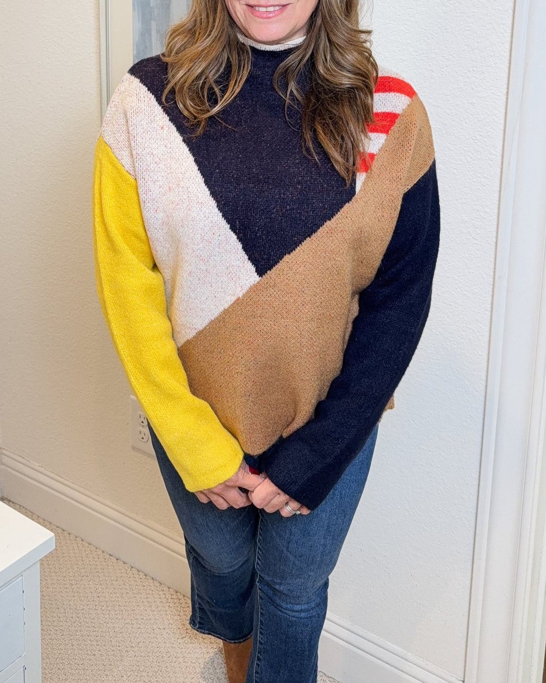 Woman wearing a colorful sweater standing in a room with a lamp and framed pictures on the wall.
