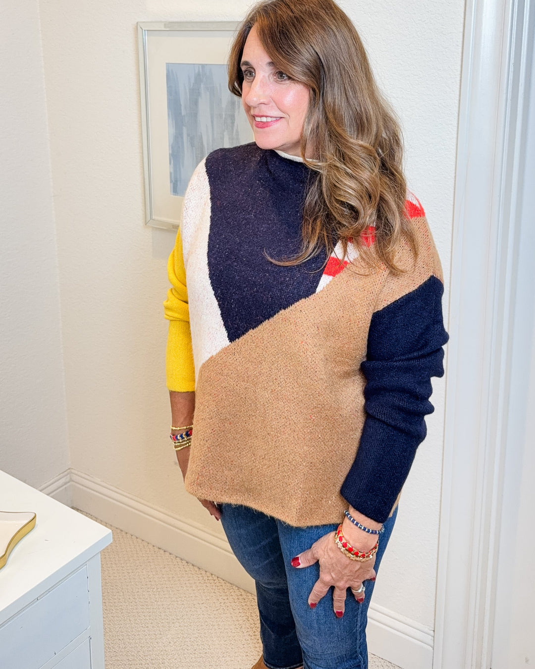 Woman wearing a colorful sweater with a beige vest in a room with framed art on the wall.