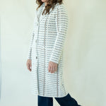 A woman modeling a long white coat with a black geometric pattern, paired with dark blue jeans and tan boots, against a plain off-white studio background.