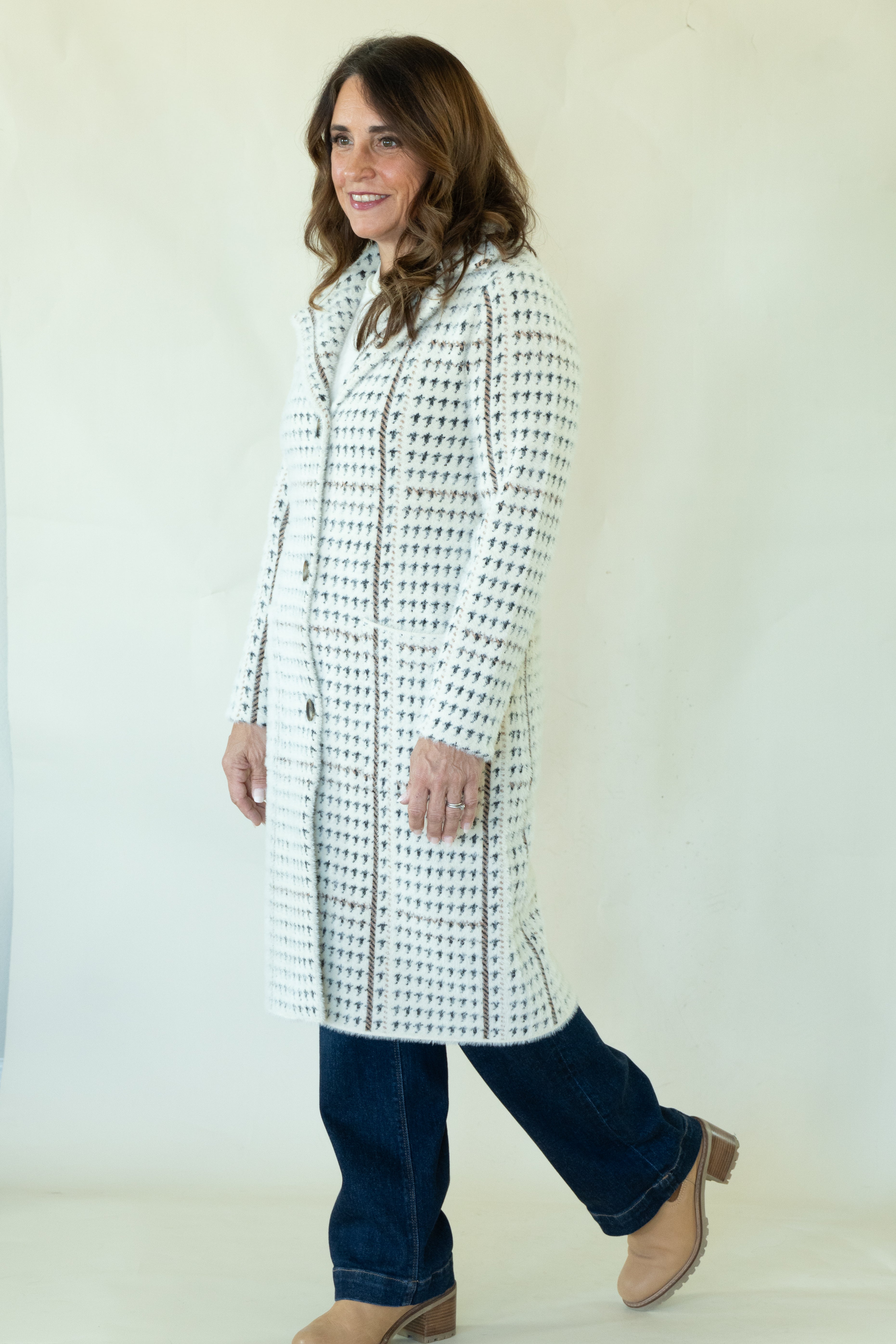 A woman modeling a long white coat with a black geometric pattern, paired with dark blue jeans and tan boots, against a plain off-white studio background.
