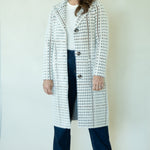 A woman modeling a long white coat with a black houndstooth pattern, worn over a white top and dark blue jeans, standing against a plain off-white background.