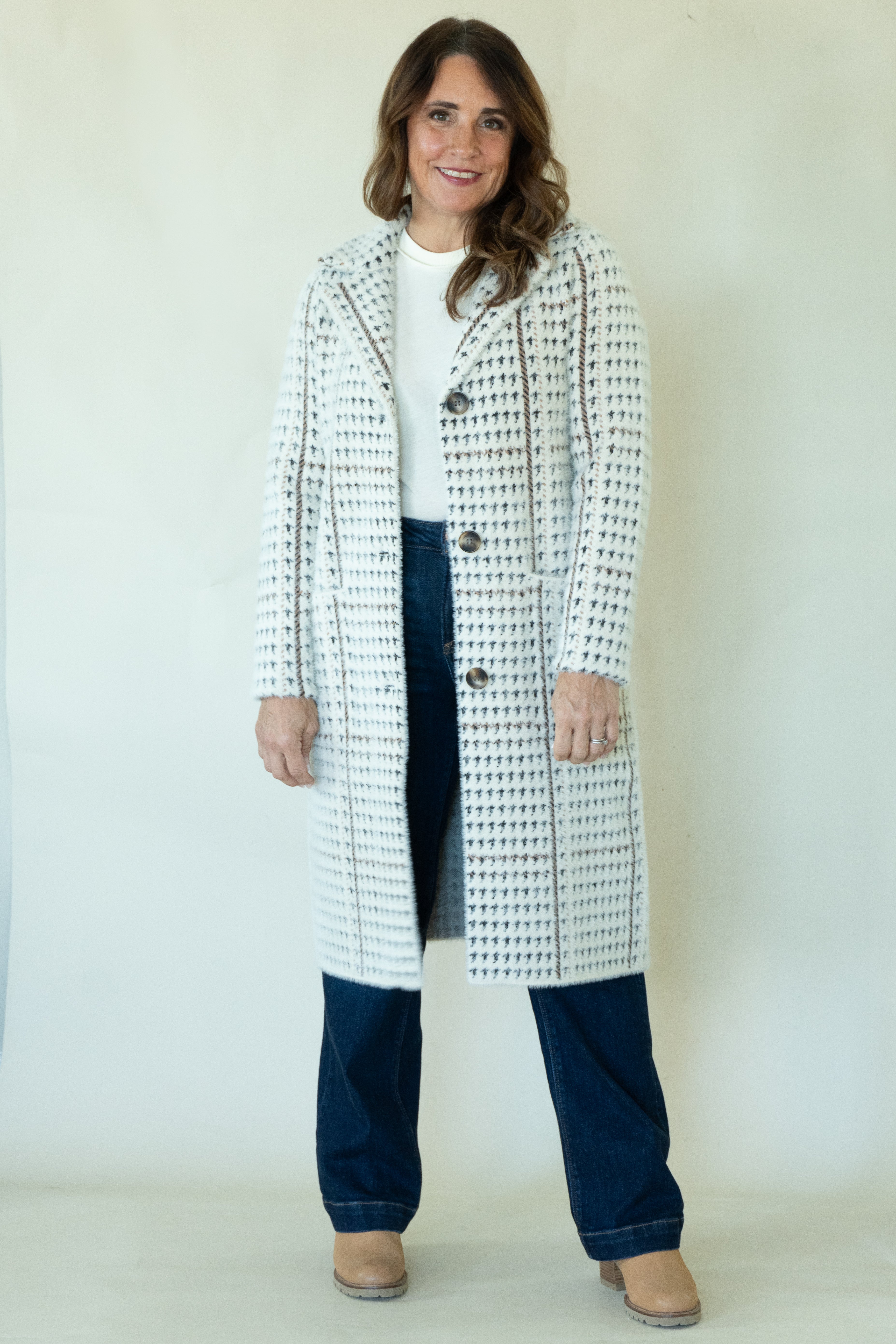 A woman modeling a long white coat with a black houndstooth pattern, worn over a white top and dark blue jeans, standing against a plain off-white background.