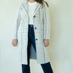 A woman modeling a long white tweed coat with black and brown houndstooth-like pattern details, worn over a white top and dark blue jeans, standing against a plain off-white background.