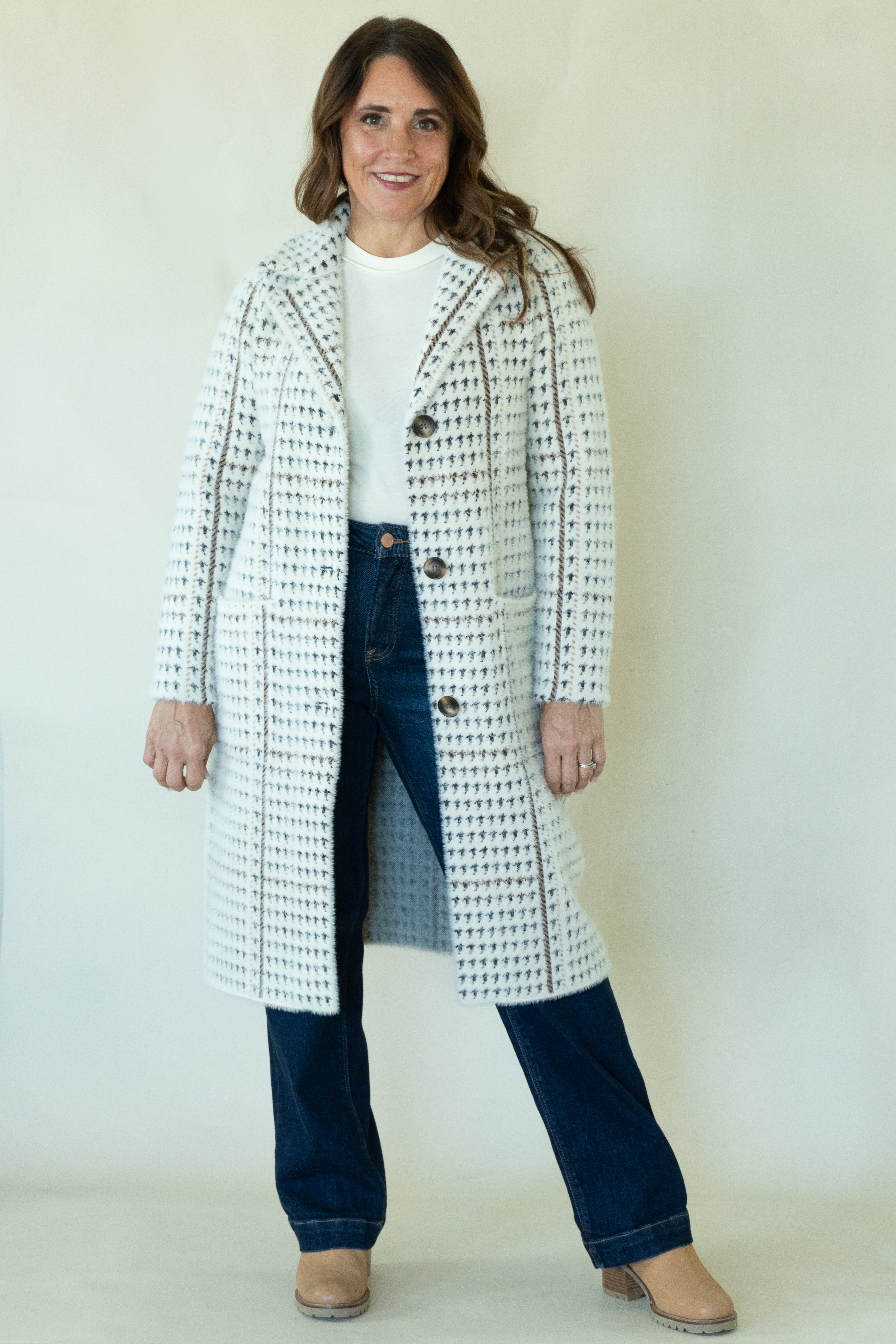 A woman modeling a long white tweed coat with black and brown houndstooth-like pattern details, worn over a white top and dark blue jeans, standing against a plain off-white background.