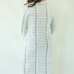 A woman viewed from behind wearing a long white knit cardigan with a black geometric lattice pattern and dark blue jeans, standing against a plain off-white wall.