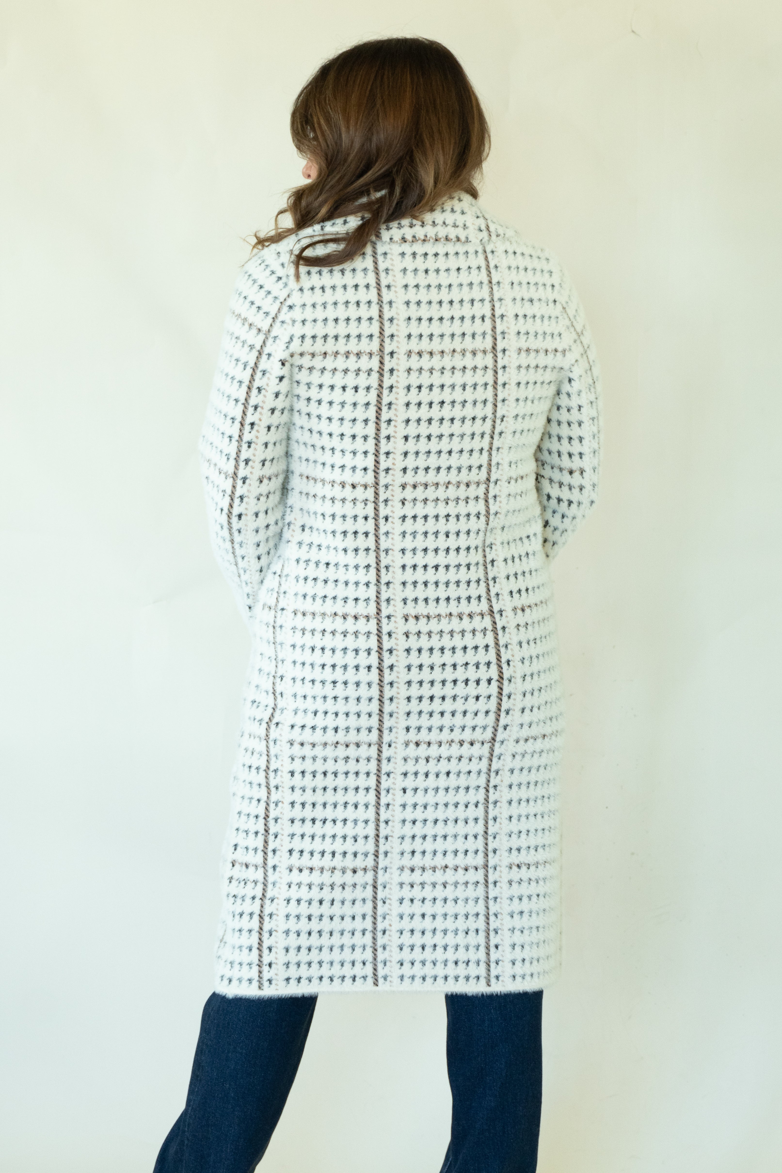 A woman viewed from behind wearing a long white knit cardigan with a black geometric lattice pattern and dark blue jeans, standing against a plain off-white wall.