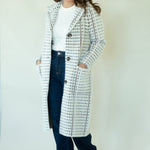 A woman modeling a long white tweed coat with a black houndstooth-like pattern, worn over a white top and dark blue jeans, standing against a plain off-white studio background.