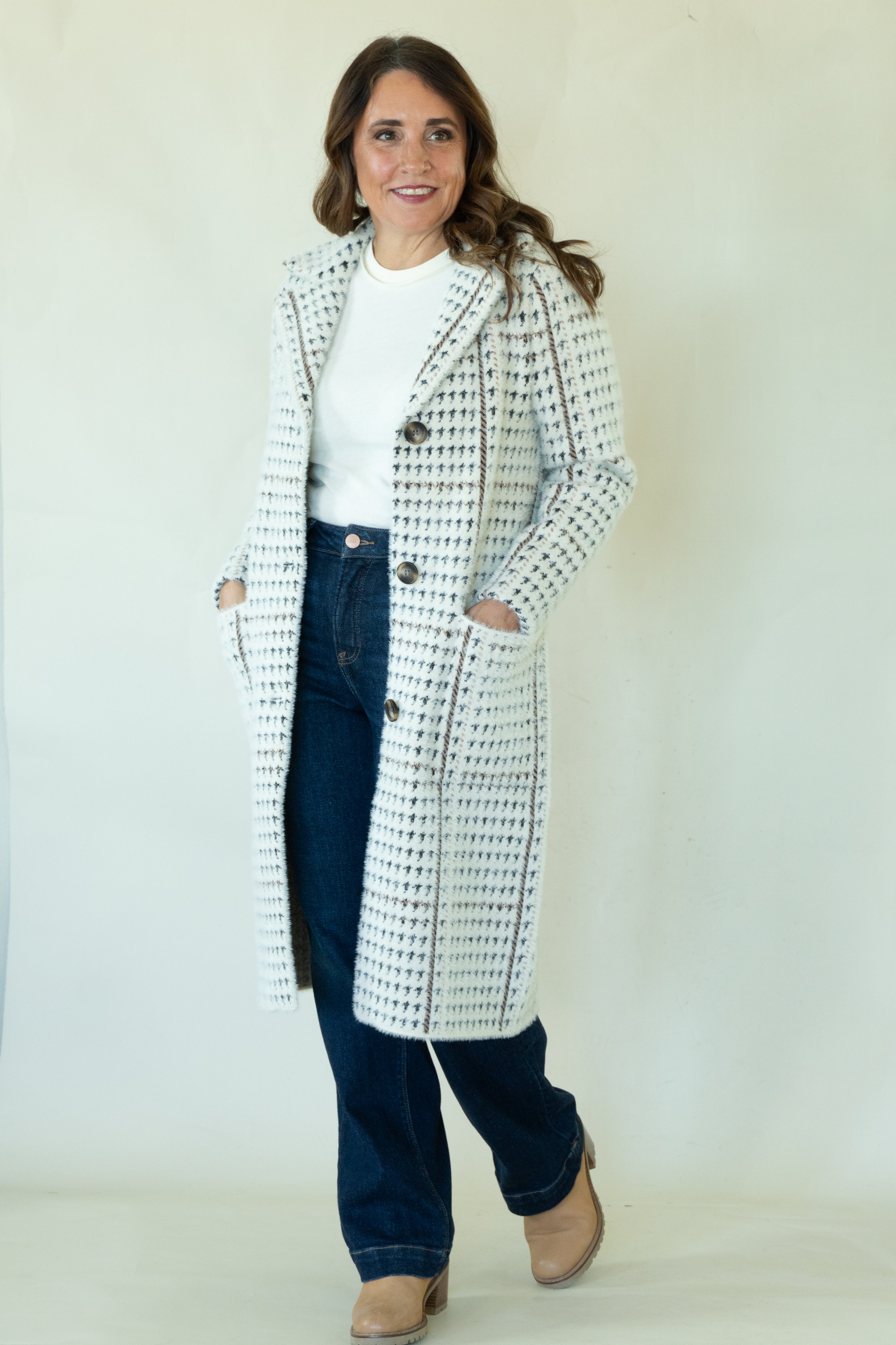 A woman modeling a long white tweed coat with a black houndstooth-like pattern, worn over a white top and dark blue jeans, standing against a plain off-white studio background.