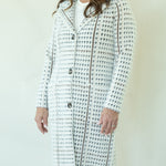 A woman wearing a white tweed coat with a black houndstooth pattern and brown trim, paired with a white top and dark blue jeans, standing against a plain off-white background.