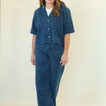 A woman modeling a matching two-piece lounge set featuring a short-sleeve button-front top and wide-leg pants, both in a dark blue with a lighter blue geometric pattern, over a white undershirt.
