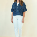 A woman modeling a short-sleeve button-up shirt with a blue and white geometric pattern, paired with white pants and beige sandals, against a plain cream background.