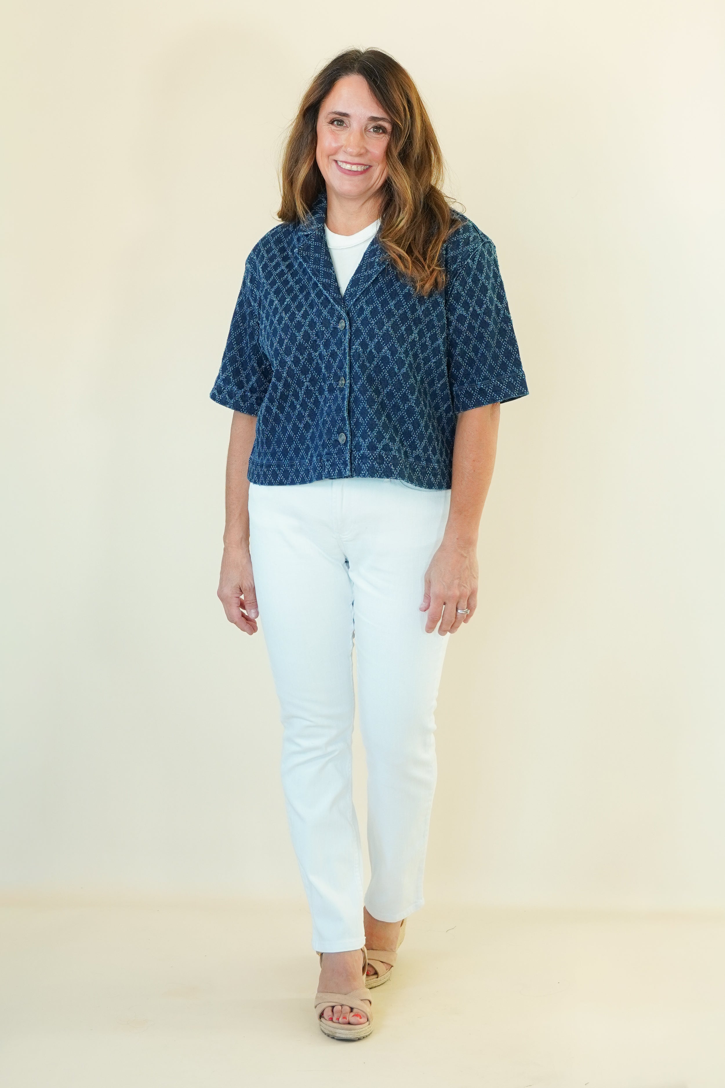 A woman modeling a short-sleeve button-up shirt with a blue and white geometric pattern, paired with white pants and beige sandals, against a plain cream background.