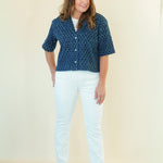 A woman modeling a short-sleeve button-up shirt with a blue and white geometric pattern, paired with white pants and beige wedge sandals, standing against a plain cream background.