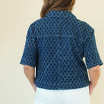 A woman shown from the back wearing a dark blue short-sleeve button-up shirt with a white geometric pattern, paired with white pants and beige wedge sandals, against a plain cream background.