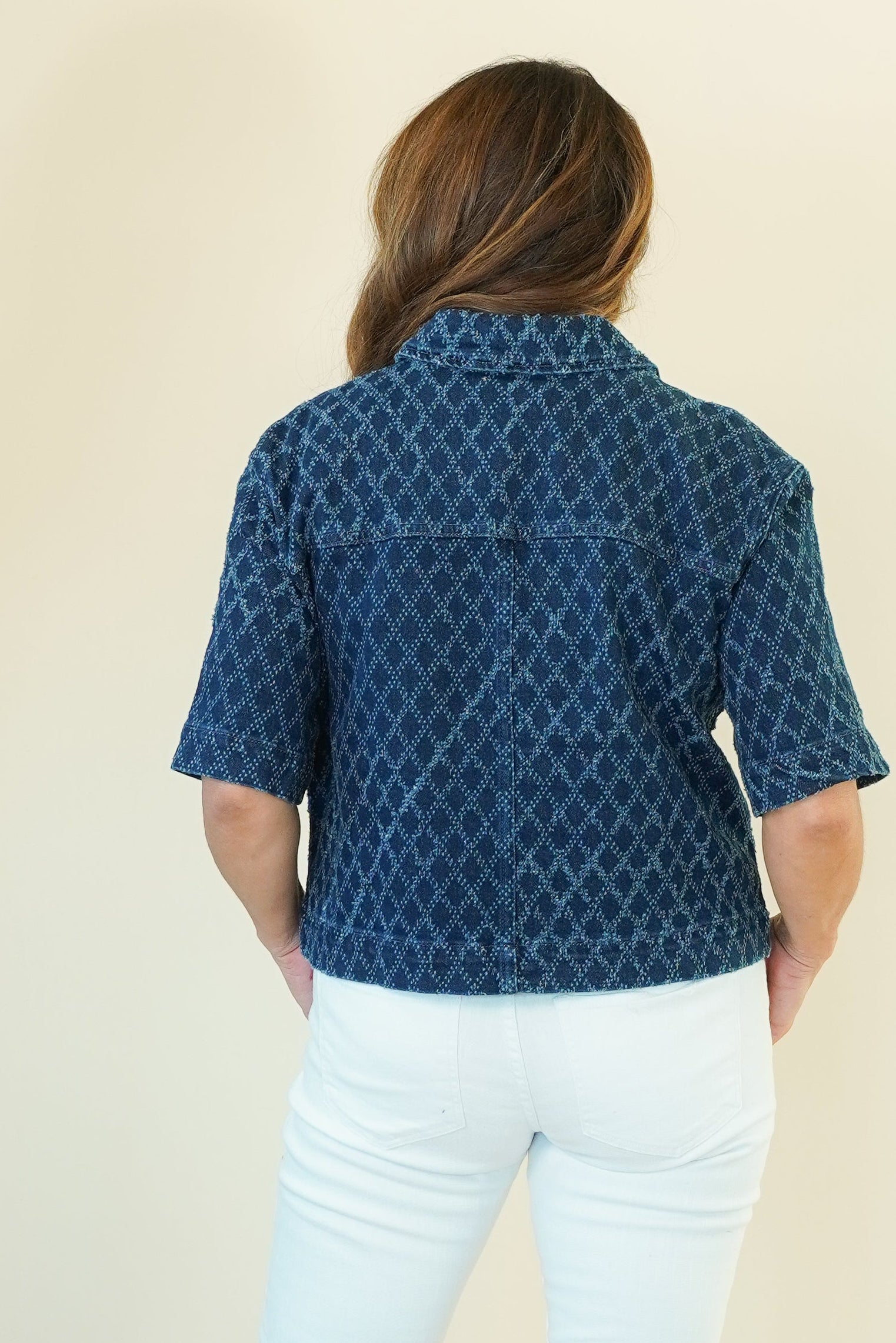 A woman shown from the back wearing a dark blue short-sleeve button-up shirt with a white geometric pattern, paired with white pants and beige wedge sandals, against a plain cream background.