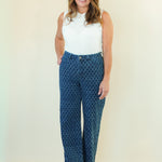 A woman modeling a pair of high-waisted, wide-leg pants with a blue and black geometric pattern, paired with a white sleeveless top, standing against a plain cream-colored background.