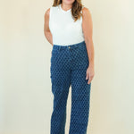 A woman modeling a pair of high-waisted, wide-leg blue pants with a white geometric pattern, paired with a white sleeveless top, against a plain cream background.