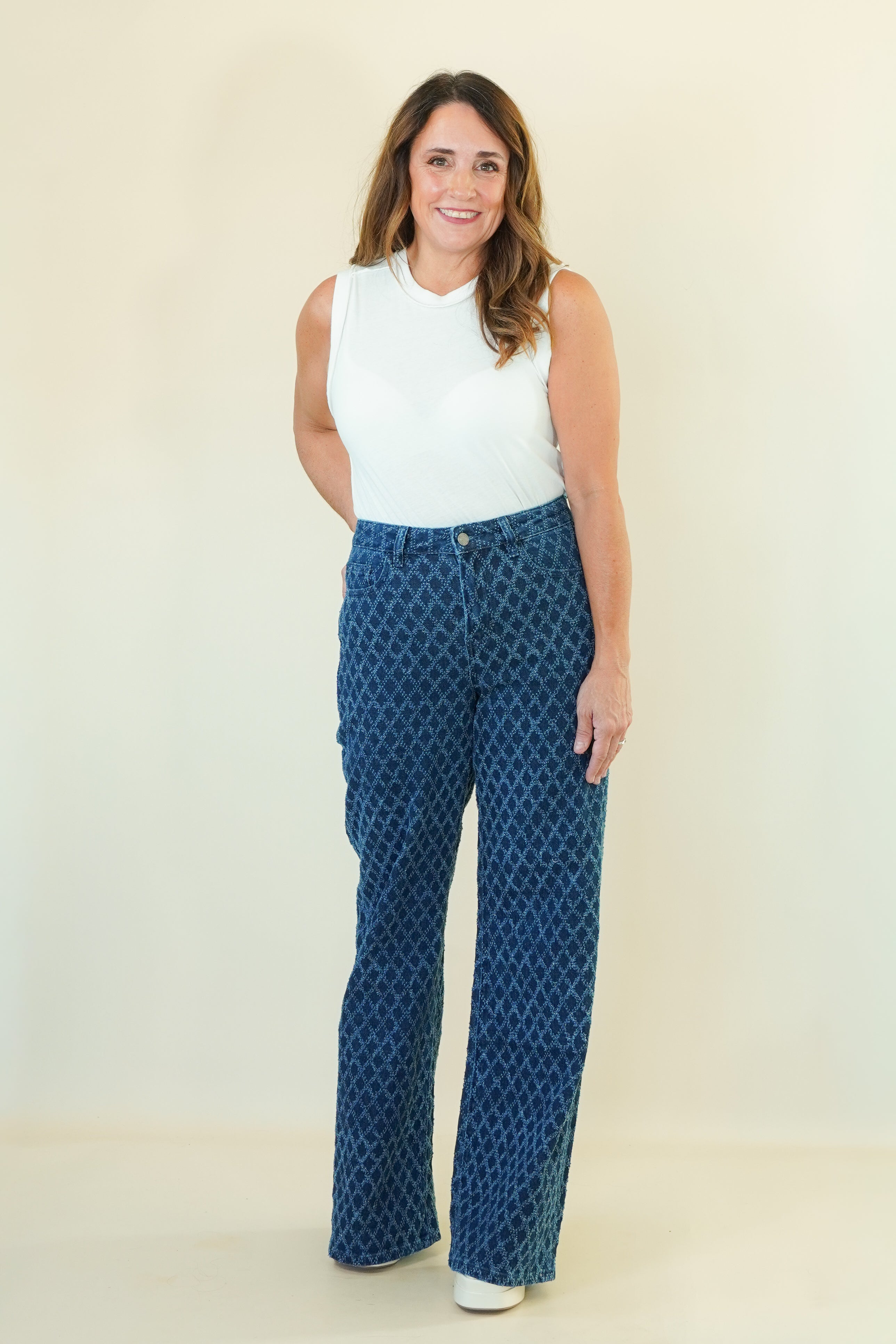 A woman modeling a pair of high-waisted, wide-leg blue pants with a white geometric pattern, paired with a white sleeveless top, against a plain cream background.