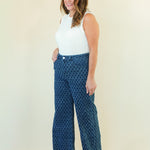 A woman modeling a pair of high-waisted, wide-leg pants with a blue and black geometric pattern, paired with a white sleeveless top, standing against a plain cream-colored background.