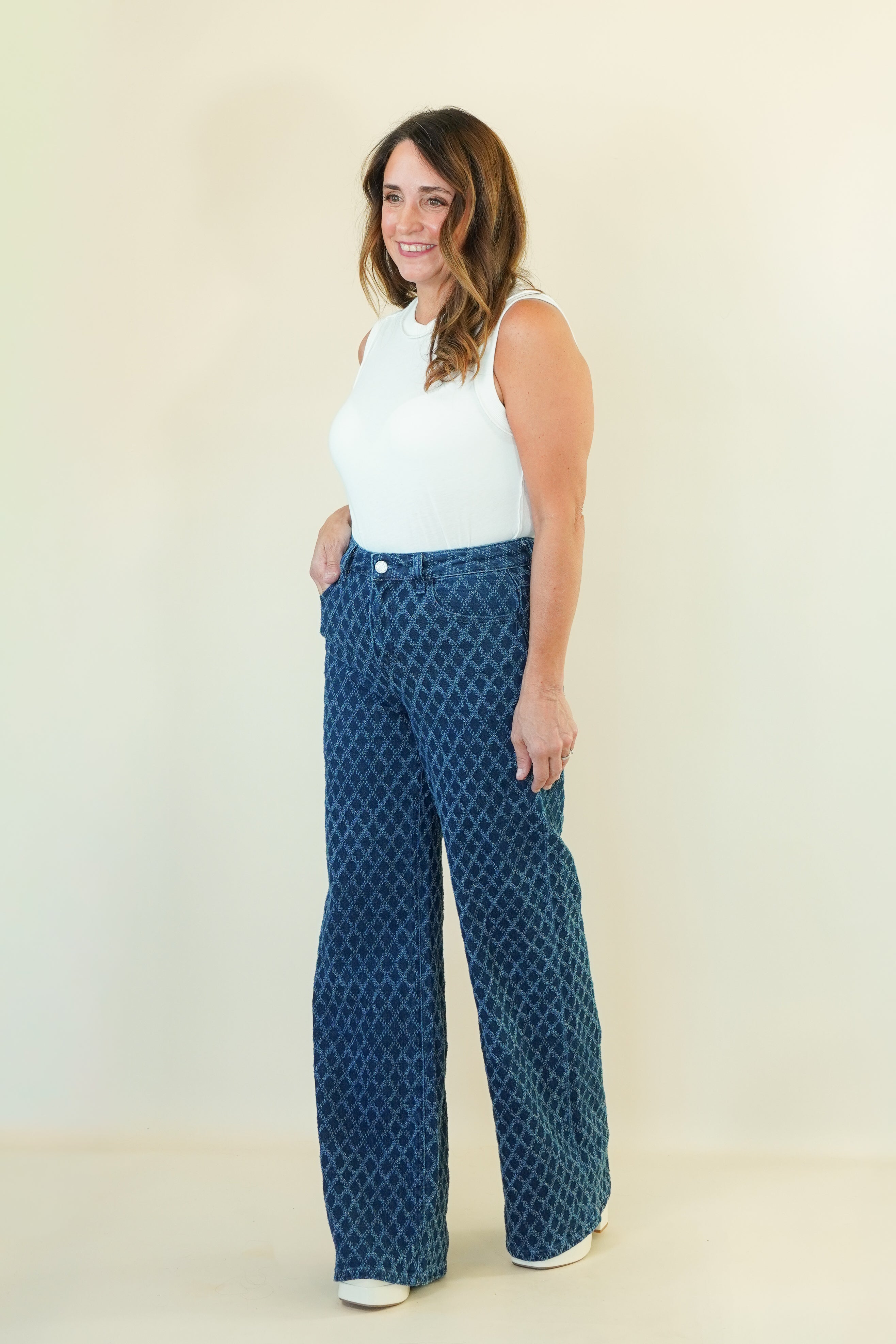 A woman modeling a pair of high-waisted, wide-leg pants with a blue and black geometric pattern, paired with a white sleeveless top, standing against a plain cream-colored background.