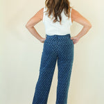 A woman shown from the back wearing a white sleeveless top and blue wide-leg pants with a repeating geometric pattern, standing against a plain cream background.