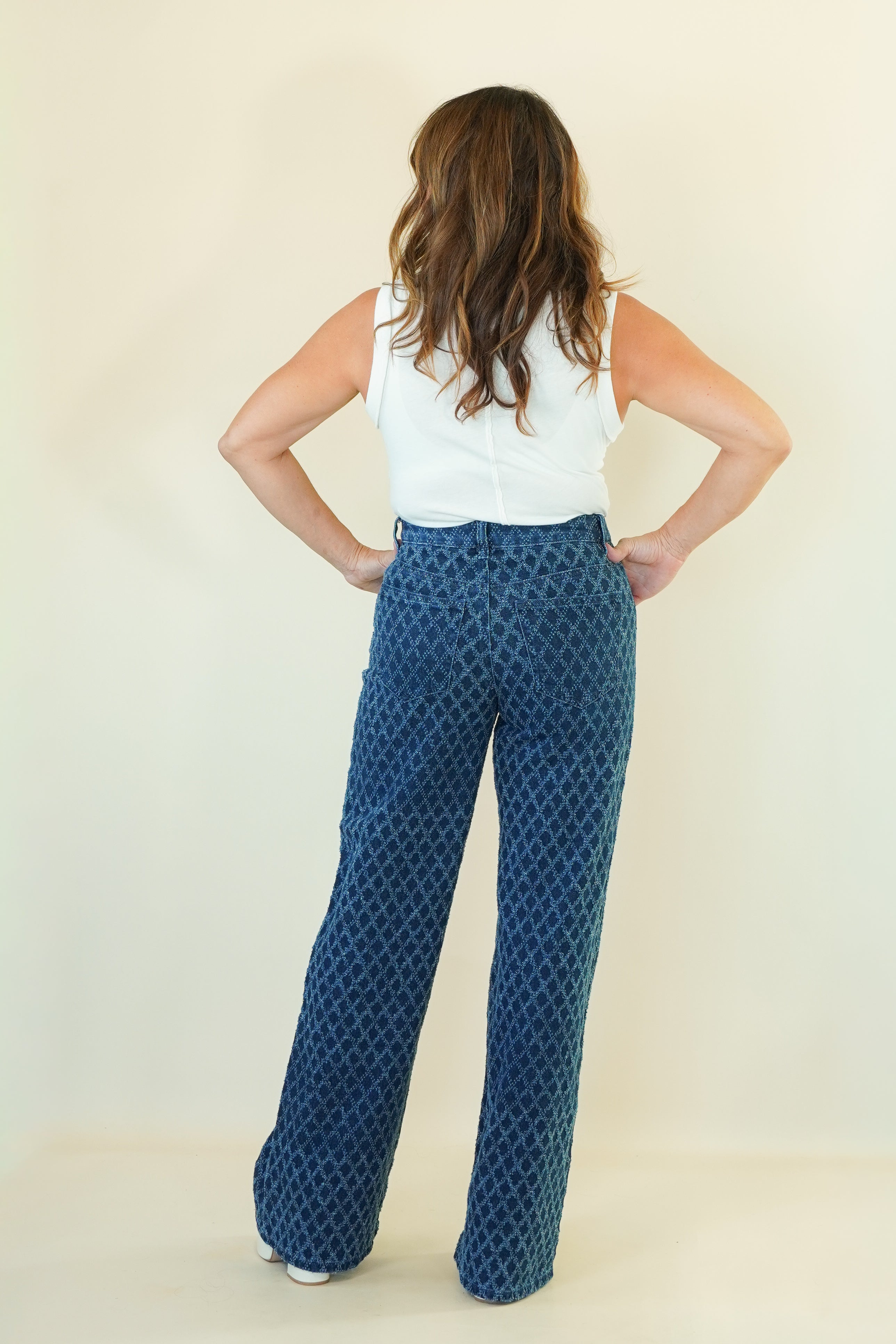 A woman shown from the back wearing a white sleeveless top and blue wide-leg pants with a repeating geometric pattern, standing against a plain cream background.