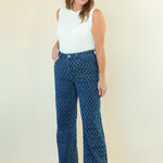 A woman modeling a pair of high-waisted, wide-leg pants with a blue and black geometric pattern, paired with a white sleeveless top, standing against a plain cream-colored background.