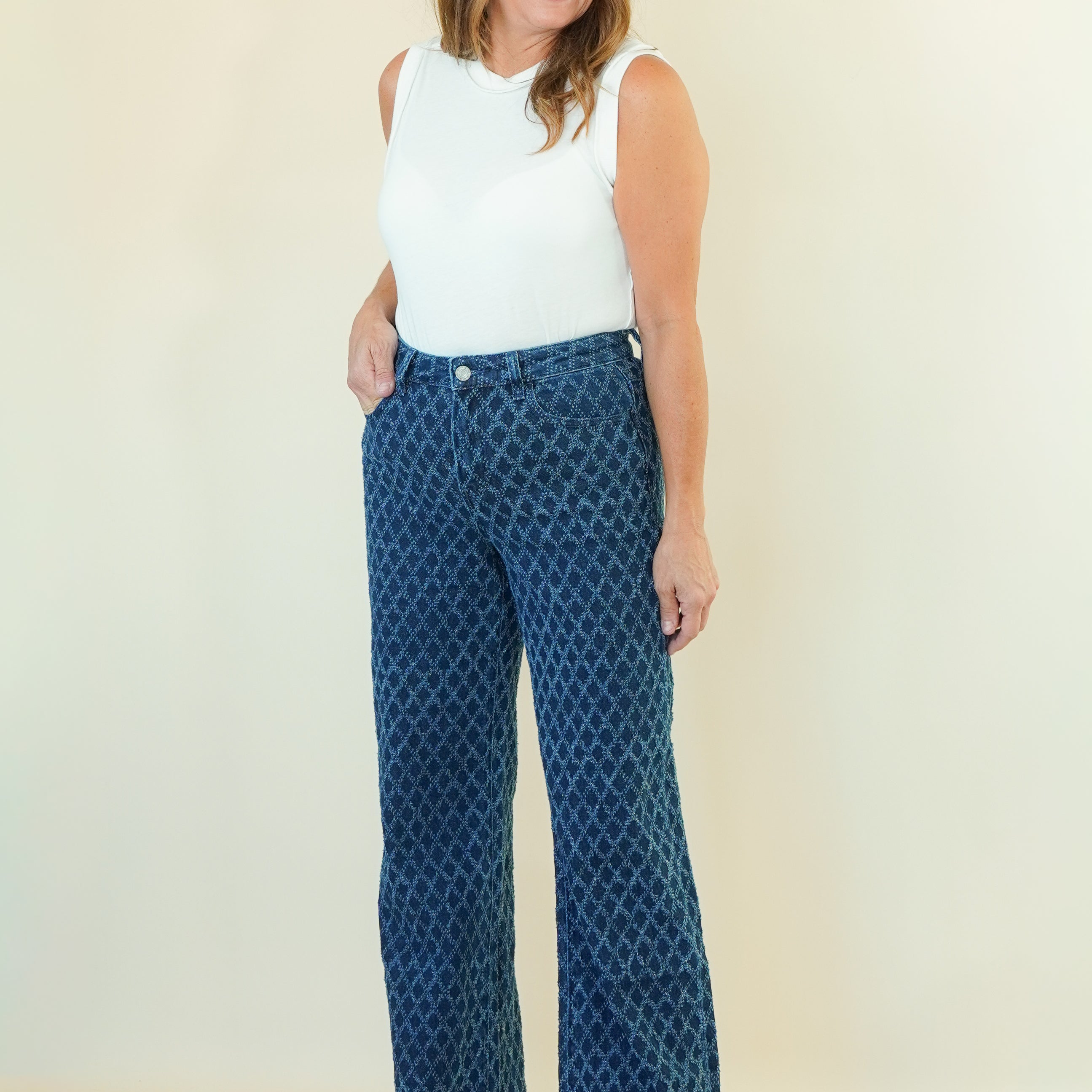 A woman modeling a pair of high-waisted, wide-leg pants with a blue and black geometric pattern, paired with a white sleeveless top, standing against a plain cream-colored background.