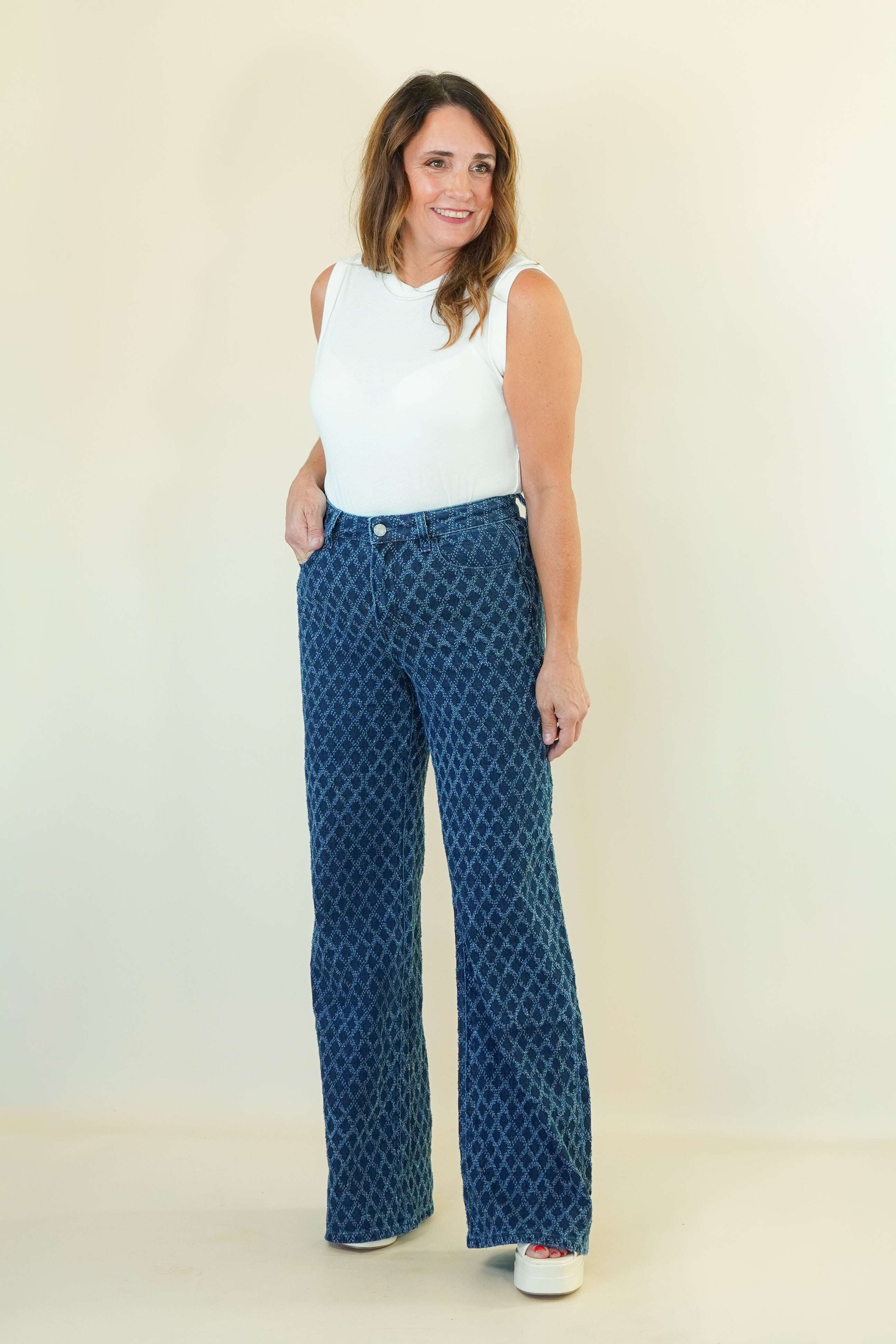 A woman modeling a pair of high-waisted, wide-leg pants with a blue and black geometric pattern, paired with a white sleeveless top, standing against a plain cream-colored background.