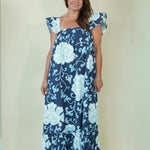 A woman modeling a navy blue maxi dress with a light blue floral pattern, featuring ruffled cap sleeves and a square neckline, set against a plain cream background.