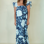 A woman modeling a navy blue maxi dress with a light blue floral print, featuring ruffled cap sleeves and a tiered ruffle hem, set against a plain cream studio background.