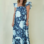 A woman modeling a navy blue maxi dress with a light blue floral pattern and ruffled cap sleeves, standing against a plain beige studio background.