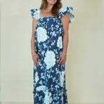 A woman modeling a navy blue maxi dress with a light blue and white floral pattern, featuring ruffled cap sleeves and a tiered ruffle hem, set against a plain beige background.