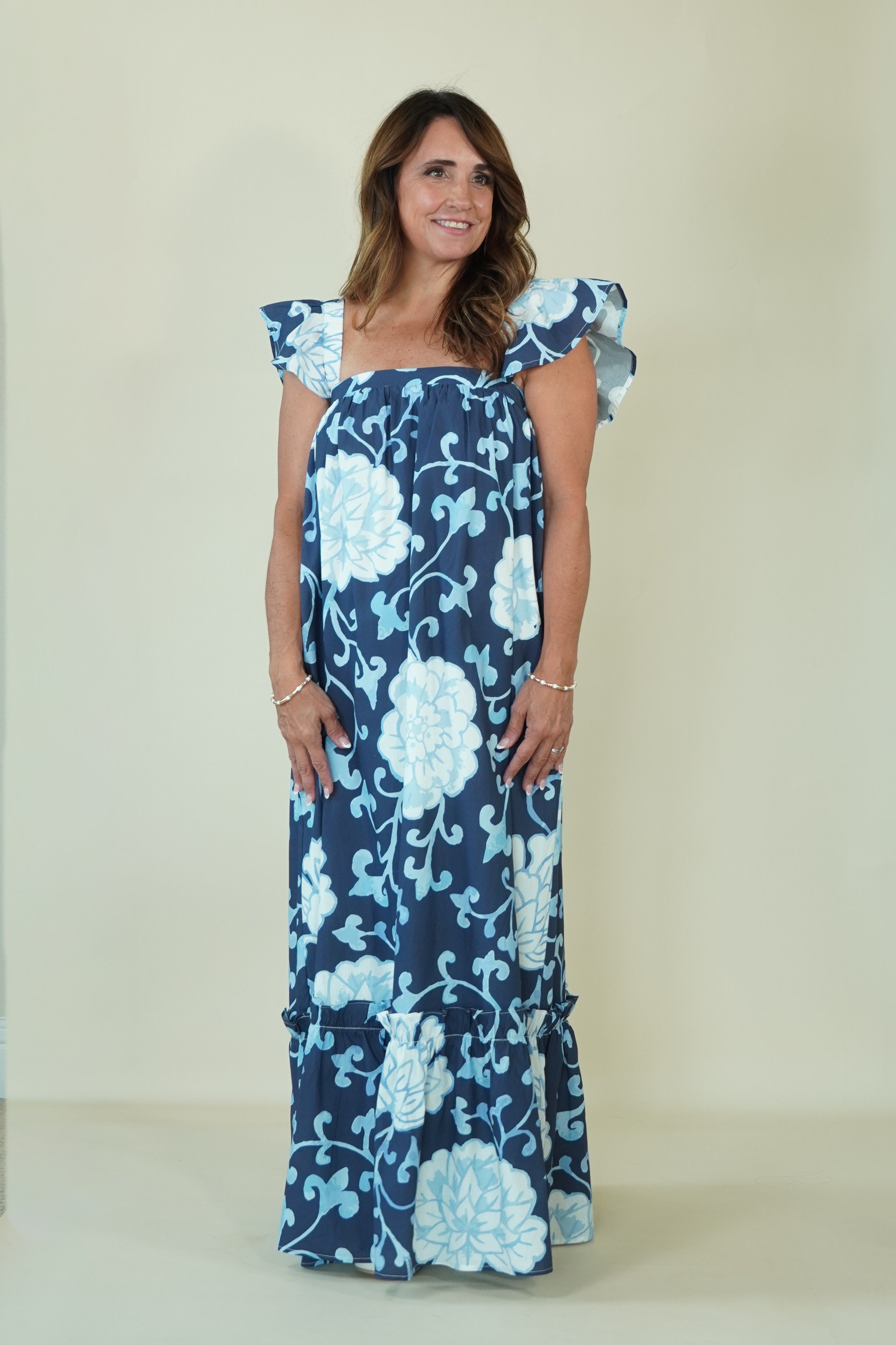 A woman modeling a navy blue maxi dress with a light blue and white floral pattern, featuring ruffled cap sleeves and a tiered ruffle hem, set against a plain beige background.