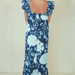 A woman viewed from the back wearing a navy maxi dress with a light blue floral print and ruffled shoulder straps, set against a plain cream background.