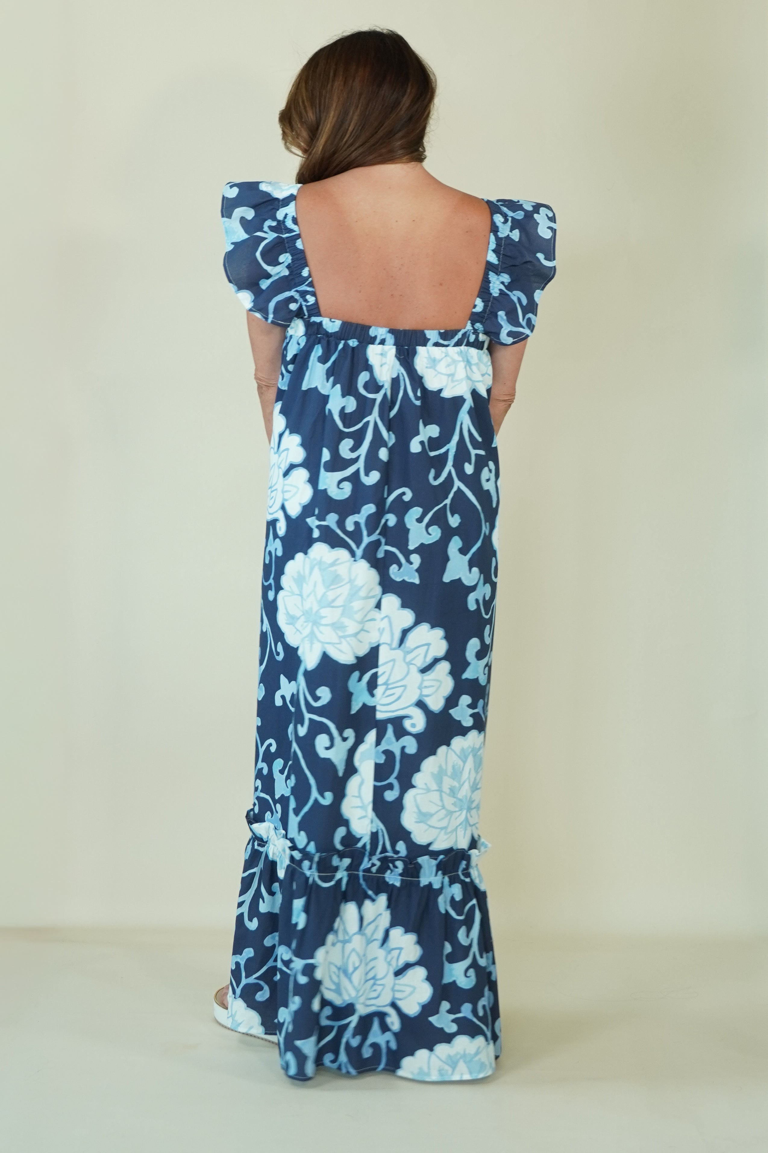 A woman viewed from the back wearing a navy maxi dress with a light blue floral print and ruffled shoulder straps, set against a plain cream background.