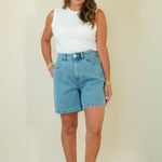 A woman modeling a white sleeveless top and light-wash blue denim shorts against a plain beige studio background.