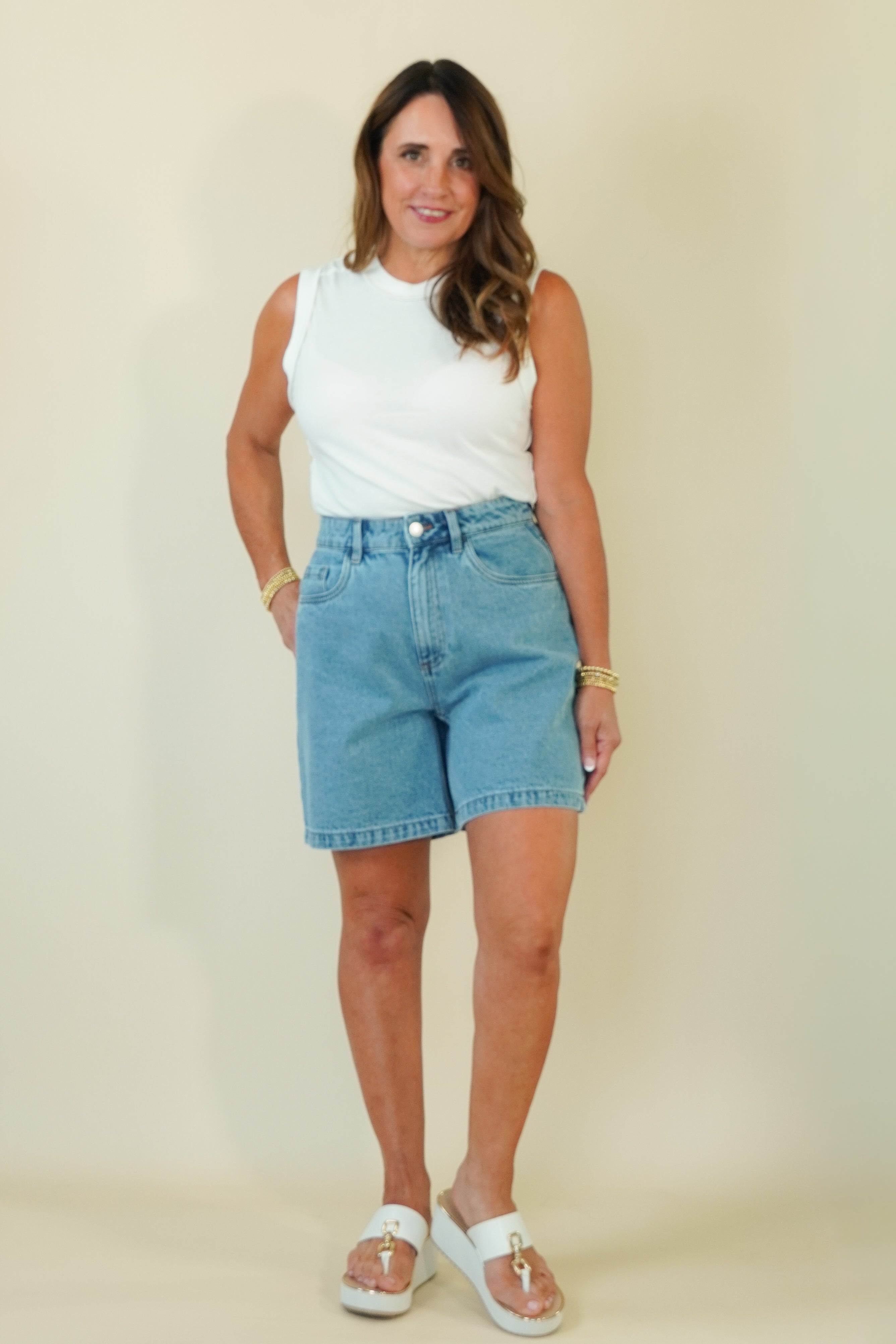 A woman modeling a white sleeveless top and light-wash blue denim shorts against a plain beige studio background.
