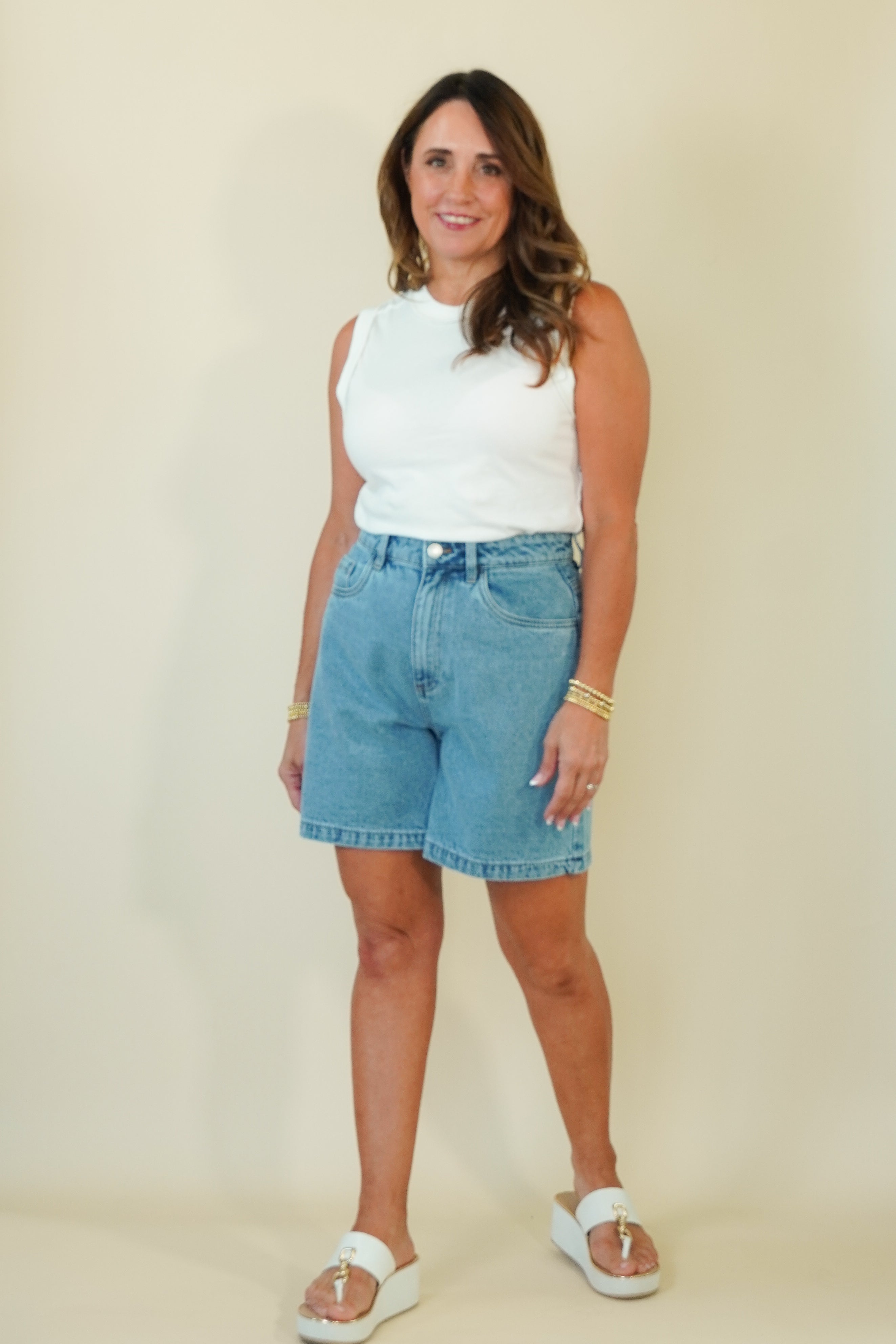 A woman modeling a white sleeveless top and light blue denim shorts against a plain beige studio background.