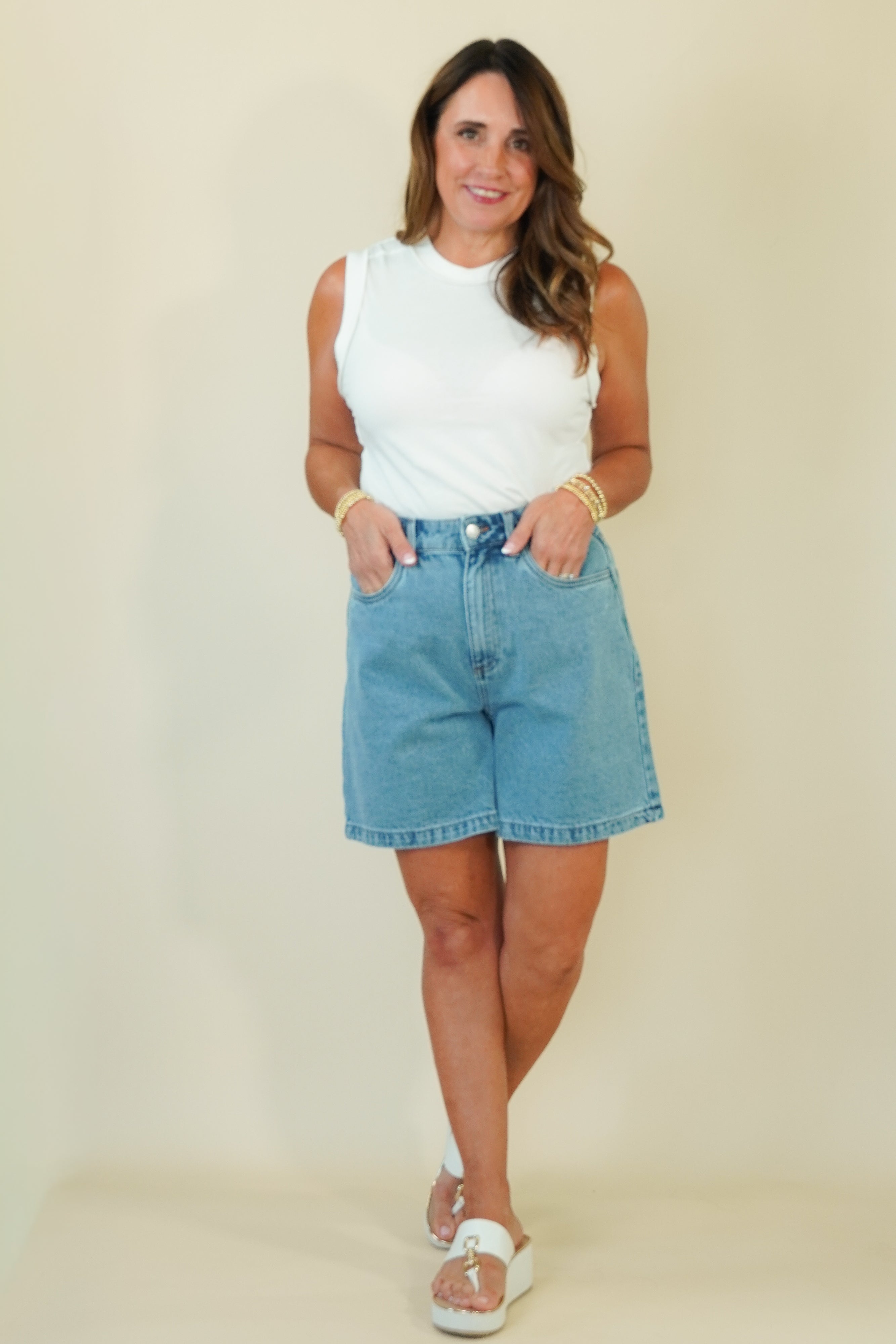 A woman modeling a white sleeveless top and light blue denim shorts against a plain beige studio background.