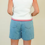 A person shown from the back wearing a sleeveless white knit top with red trim and light blue denim shorts against a plain beige studio background.