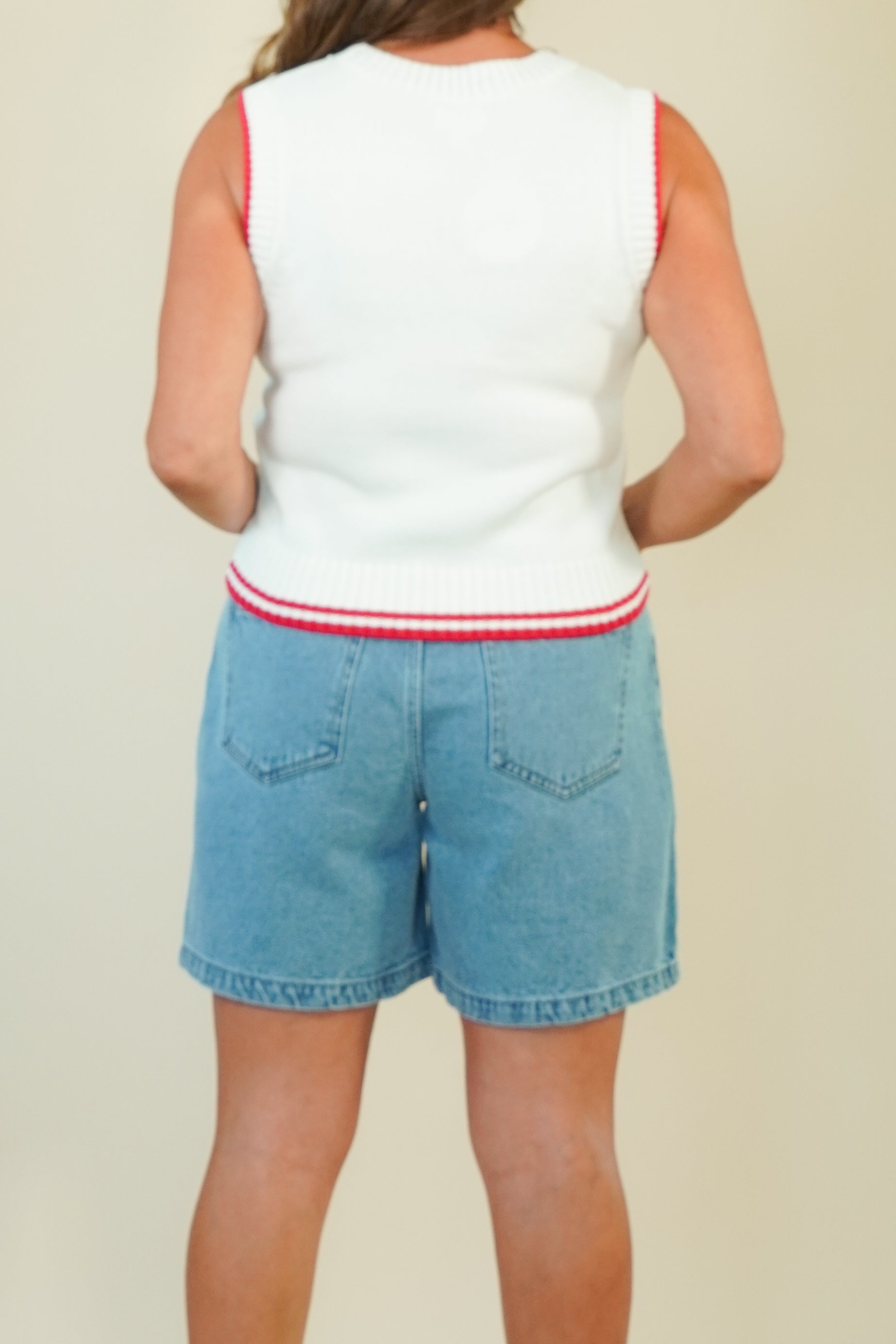 A person shown from the back wearing a sleeveless white knit top with red trim and light blue denim shorts against a plain beige studio background.