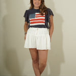Woman wearing a t-shirt with an American flag design and white shorts against a plain background